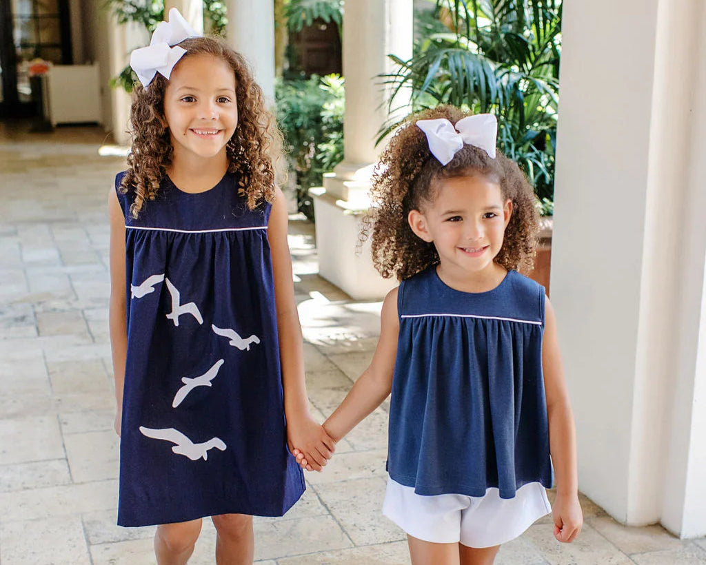 Two young girls in matching navy dresses with white bird patterns, standing in a sunlit outdoor setting.
